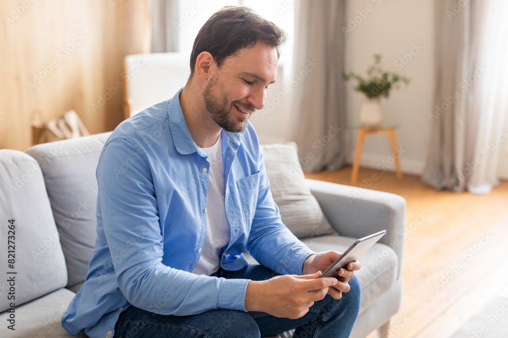 Poster A man is comfortably seated on a plush couch, engrossed in using a modern tablet device. He is focused on the screen, tapping and scrolling with his fingers as he interacts with the digital interface.