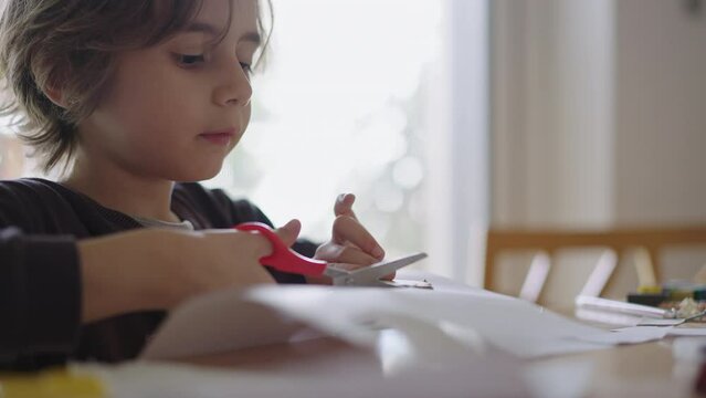 A young boy concentrates on a paper craft project while referring to instructions on a digital tablet at a table in a well-lit room.