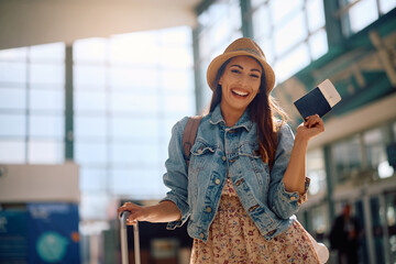 Happy woman holding her passport and plane ticket at departure area and looking at camera.