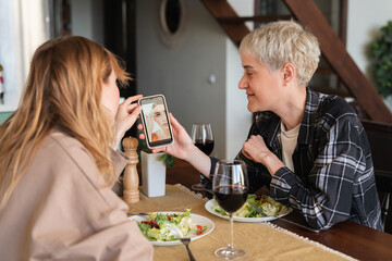 Two young girlfriends lesbians showing photos on smartphone during romantic date dinner discussing skin care problems and beauty tips. Best friends, LGBT relationship and friendship