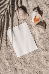 Blank paper card with mockup copy space and sea shells on beach sand texture with palm tree foliage...