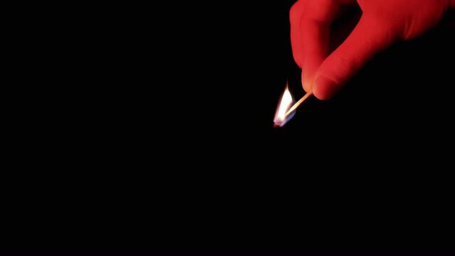 Hand Holds a Burning Match in Red Lighting on a Black Background in Empty Space. Side view. One burning match in the hand burns with a bright flame. Close-up, slow motion. Isolated. Dark room.