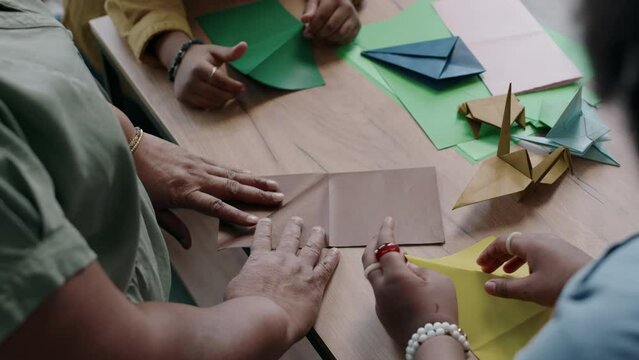 Close-up of dark-skinned hands of unrecognizable children and woman carefully folding colour origami paper into various shapes and crane figures