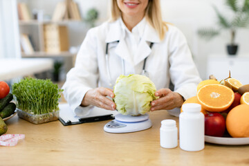 Cropped view of hands of female nutritionist sitting at table in clinic and weighs cabbage, making...