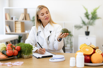 Mature Caucasian female nutritionist making healthy eating plan and calculating calorie content of avocado using weight. Happy female writing prescription for patients proper healthy diet.