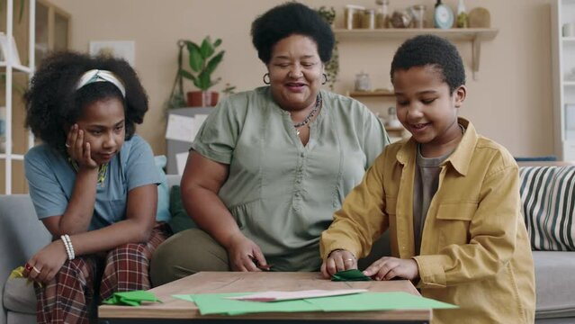 Medium of African American children cheerfully playing with origami paper frog, making it jump on table, showing handiwork to grandmother, sitting on couch in cozy home