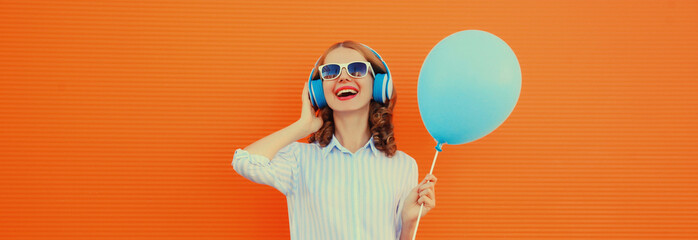 happy cheerful young woman in headphones listening to music with blue balloon on orange background