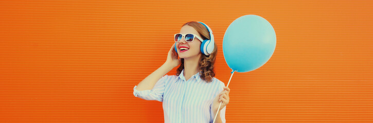 happy cheerful young woman in headphones listening to music with blue balloon on orange background