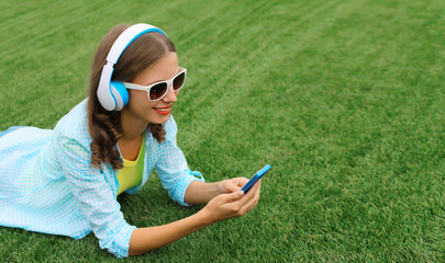 Happy young woman holding phone listening to music in headphones lying on grass in summer park