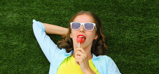 Happy teenager girl resting with lollipop lying on green grass in summer park