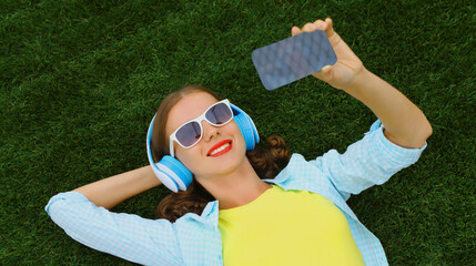 Happy young woman taking selfie with smartphone listening to music lying on grass in summer park