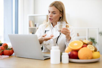 Female doctor in white coat holding glass of water during a video call. Experienced female nutritionist tells patient about benefits of water during an online session using laptop.