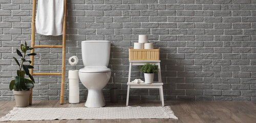 Interior of restroom with toilet bowl, ladder with towel and toilet paper near grey brick wall