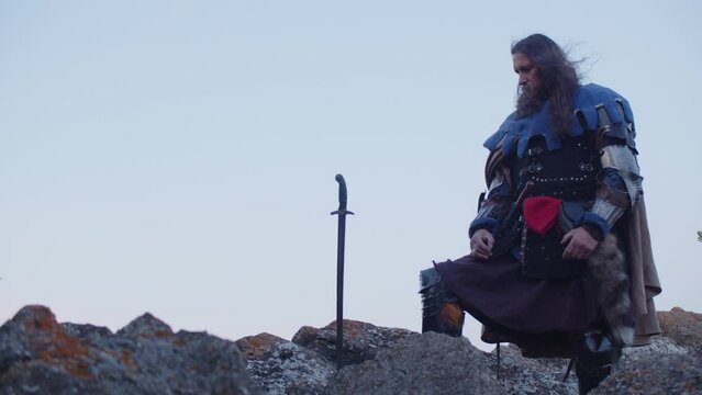 A man in a medieval knight costume pulls a sword from a stone against the background of a blue evening sky. Historical reconstruction. Costume performance. King Arthur