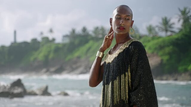 Outrageous gay black man in luxury gown poses on scenic ocean beach. Gender fluid ethnic fashion model in long posh dress and accessories looks at camera and touches neck gracefully slow motion