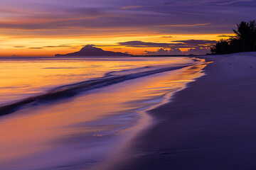 A tropical beach at dusk, with smooth white sand and calm water, the sky ablaze with shades of orange and purple, and a silhouette of a distant island