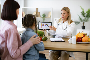 Little school girl sitting on mothers lap with visit to woman pediatrician nutritionist. Mature Caucasian female doctor shows on tablet graphs of food for healthy eating.