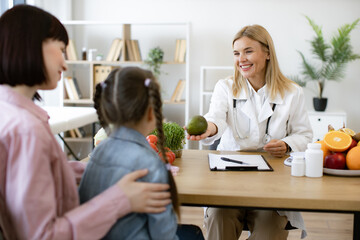Mature woman with fresh avocado in hand chatting with little female child and her mother in consulting room. Experienced medical specialist giving advice on improving diet by abundance of nutrients.