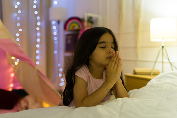 Little girl shutting her eyes and folding her hands in prayer in her cozy, warmly lit bedroom at...