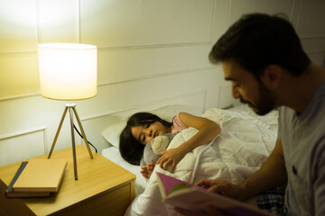 Dad reads a bedtime story to his little girl, tucked into bed at home with soft, warm lighting...
