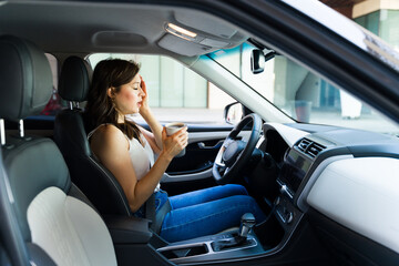 Sleepy Caucasian woman in a car and trying to wake up with some coffee in the morning while driving to work