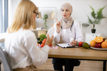 Mature Caucasian lady receives consultation on healthy eating. Muslim woman nutritionist makes plan...