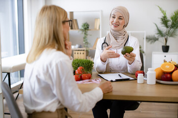 Arabian woman in hijab with fresh avocado in hand chatting with mature female lady in consulting room. Experienced medical specialist giving advice on improving diet by abundance of nutrients.