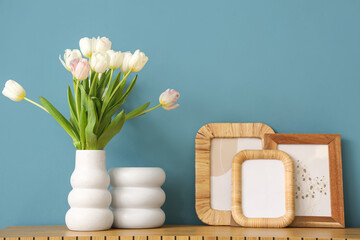 Photo frames and vase with tulips on commode near blue wall