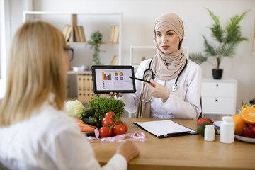 Mature lady patient visiting arabian woman nutritionist in hijab at modern clinic. Muslim female...