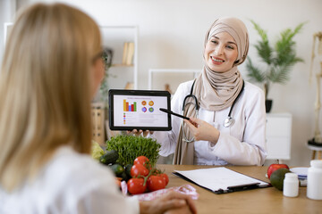Mature lady patient visiting arabian woman nutritionist in hijab at modern clinic. Muslim female doctor showing on tablet graphs of food for healthy eating.