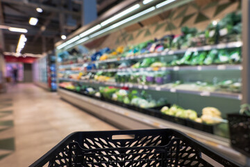Blurred image of a supermarket shopping area. A rack with fresh vegetables, fruits and herbs is...