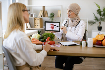 Muslim experienced female doctor showing tablet with gastrointestinal tract image for treatment planning to mature lady. Beautiful adult woman at appointment with family nutritionist.