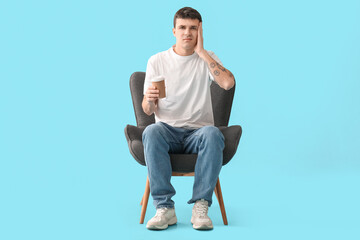 Tired young man with cup of coffee sitting in armchair on blue background