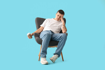 Tired young man with cup of coffee sitting in armchair on blue background
