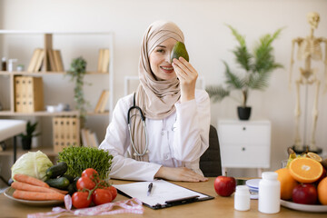 Happy female writing prescription for patients proper healthy diet. Arabian female nutritionist making healthy eating plan and calculating calorie content of avocado using weight.
