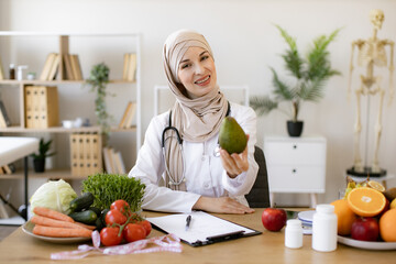 Arabian female nutritionist making healthy eating plan and calculating calorie content of avocado using weight. Happy female writing prescription for patients proper healthy diet.