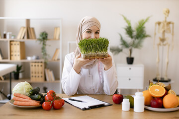 Close up view of attractive lady in white coat holding container with microgreens while sitting at desk in workplace. Muslim specialist in nutrition with hijab proposing modern addition to diet.