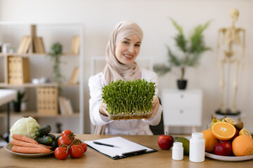 Muslim specialist in nutrition with hijab proposing modern addition to diet. Close up view of attractive lady in white coat holding container with microgreens while sitting at desk in workplace.