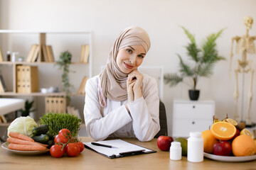 Arabian nutritionist doctor with fruits and vegetables in office. Nutritionist working writing diet plan for patient. Portrait of female nutritionist in office. Smiling young dietician sitting at desk