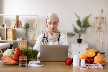 Smiling young dietician sitting at desk. Nutritionist working on laptop and writing diet plan for patient. Muslim nutritionist doctor with fruits and vegetables in office.