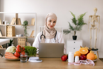 Muslim nutritionist doctor with fruits and vegetables in office. Nutritionist working on laptop and writing diet plan for patient. Smiling young dietician sitting at desk.