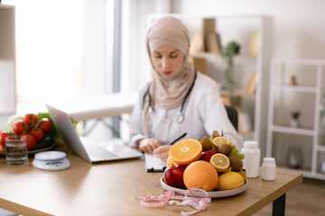 Serious Muslim lady in doctor's coat typing on modern laptop in consulting room of medical center. Experienced nutrition professional searching for weight loss information on food websites.