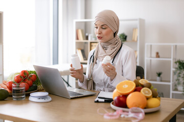 Positive arabian nutritionist woman holding pills or vitamins in hands during online consultation with patient using laptop. Muslim doctor in hijab distantly describes the treatment plan.