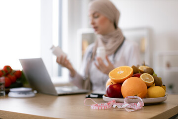Close up on fruits with measuring tape. Muslim doctor distantly describes the treatment plan. Arabian nutritionist woman holding pills or vitamins in hands during online consultation with patient.