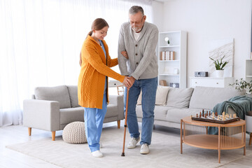 Female caregiver helping senior man with walking stick in nursing home