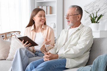 Female caregiver with senior man reading book on sofa in nursing home