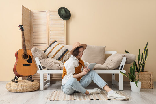Young woman in hat and eyeglasses using tablet and sitting on floor in living room - Powered by Adobe