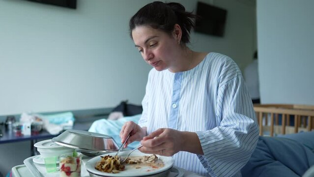 Woman in a hospital gown eating a meal on a tray while sitting on a hospital bed, showing the care and support provided to patients during their recovery period in the hospital