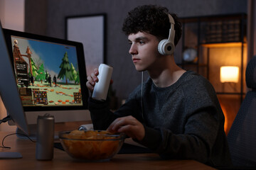 Young man with energy drink and headphones playing video game at wooden desk indoors