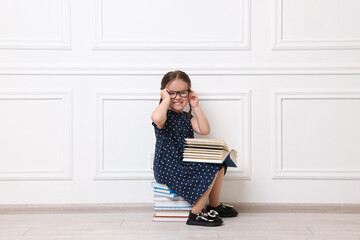 Cute little girl in glasses sitting on stack of books near white wall